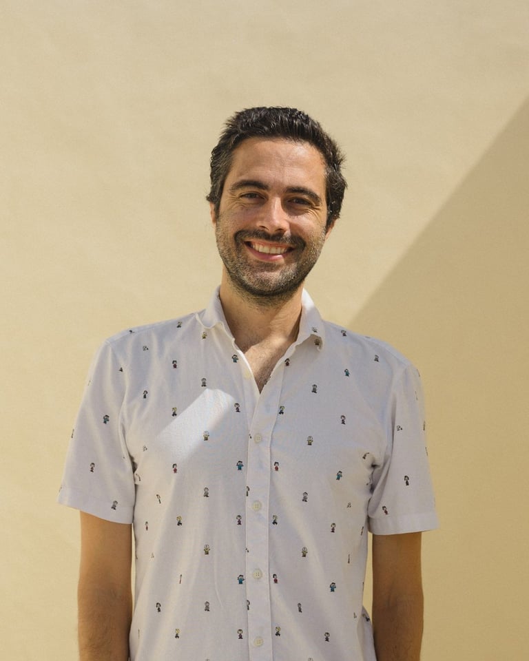 Man with dark hair and beard smiling at camera wearing light blue patterned short-sleeve shirt against beige wall