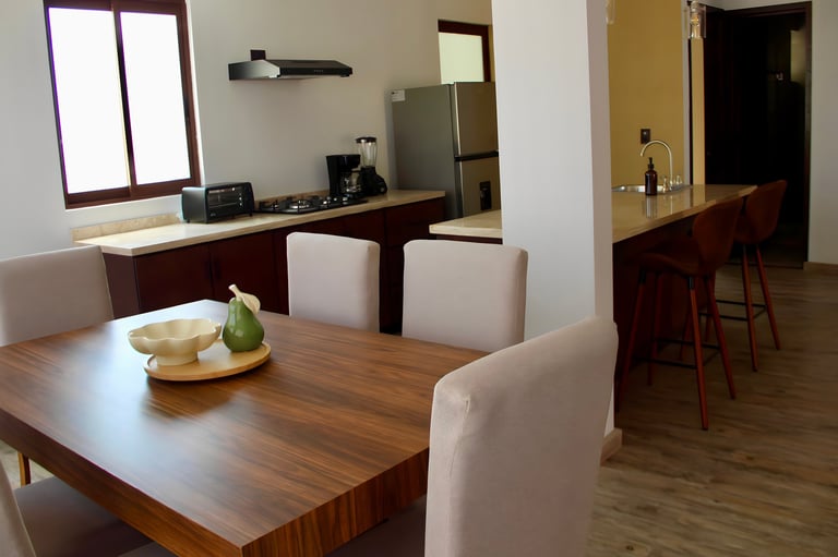 Modern kitchen and dining area with wooden table, white chairs, stainless steel appliances, and natural light from window