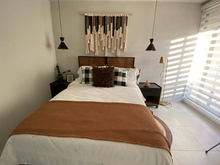 Modern bedroom with queen bed, brown and white bedding, wooden headboard, pendant lights, and white shuttered windows
