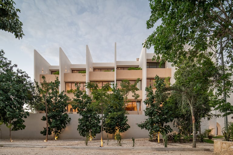 Modern terracotta building with distinctive pointed roof peaks, surrounded by mature trees in an arid landscape