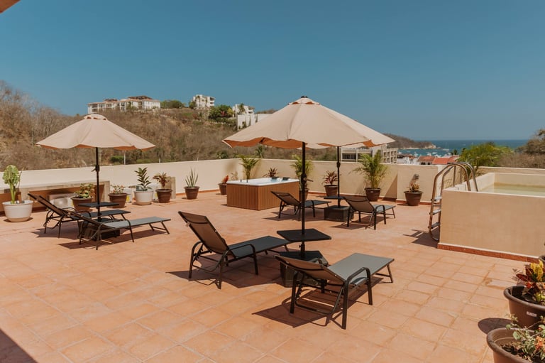Rooftop terrace with lounge chairs, beige umbrellas, potted plants, and mountain views under clear blue sky