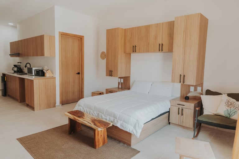 Modern bedroom with natural wood furniture, white bedding, built-in cabinetry, kitchenette area, and neutral tones throughout