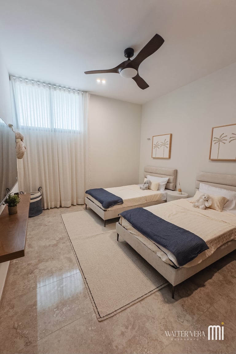 Modern bedroom with two beds, ceiling fan, white curtains, framed wall art, and neutral beige decor