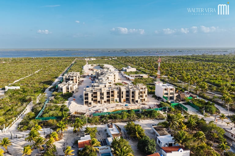 Aerial view of a modern residential complex surrounded by lush vegetation and palm trees, with a lagoon visible in the distance