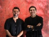 Two men in black shirts posing against a red textured background, standing side by side with relaxed postures