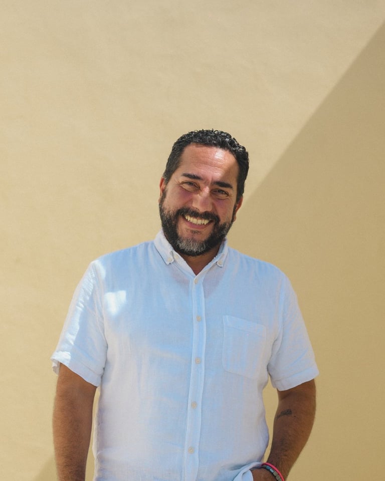 Man with dark beard and short dark hair wearing a light blue button-up shirt, smiling at camera against a beige wall