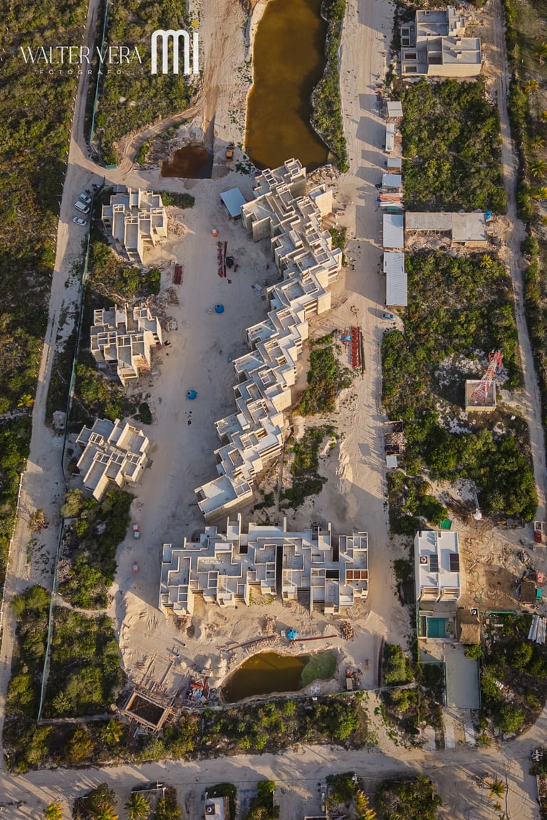 Aerial view of residential construction site with completed townhouses and ongoing building work surrounded by green vegetation and roads.