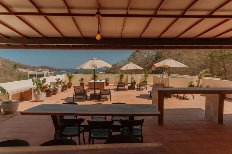 Modern terrace with wooden tables, dining chairs, beige umbrellas, and lounge seating under a timber pergola overlooking a desert landscape