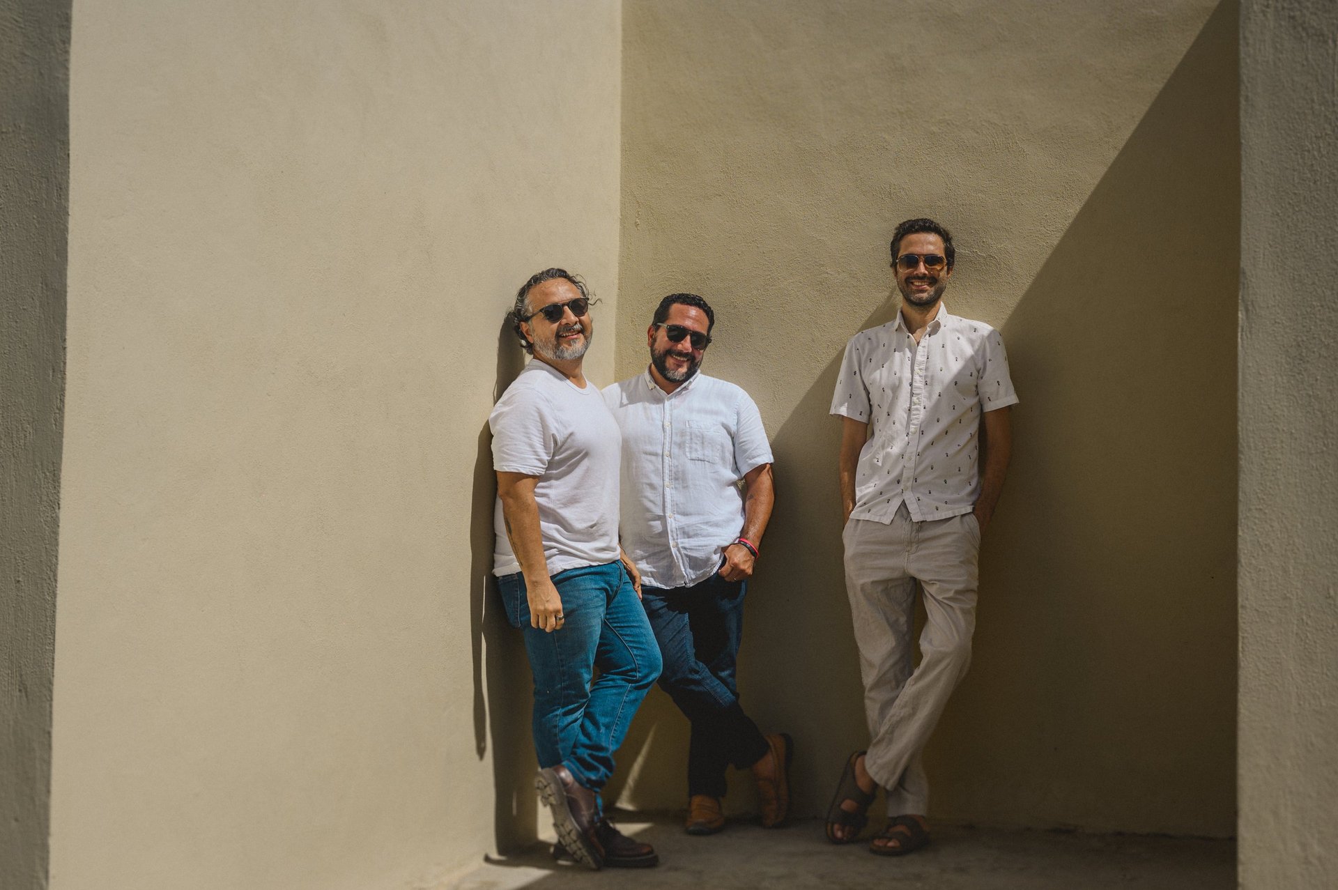 Three people wearing sunglasses and casual clothing stand together in a minimalist concrete alcove