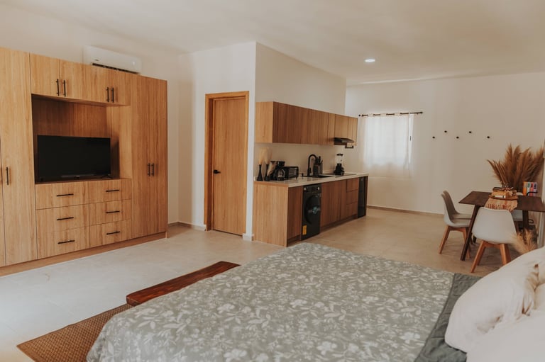 Modern studio apartment with wood cabinetry, bed in foreground, kitchenette, TV unit, and dining area with natural lighting