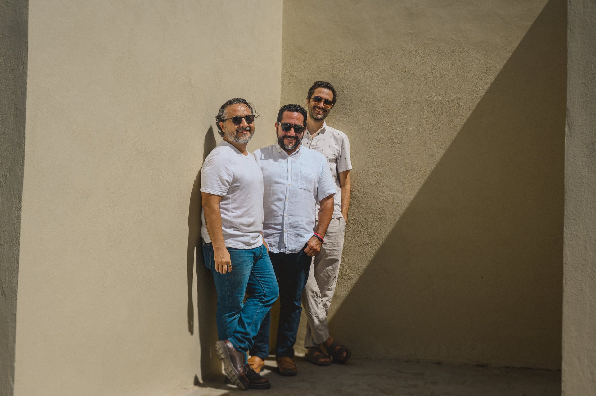 Three men in white shirts and sunglasses standing against a minimalist beige concrete wall