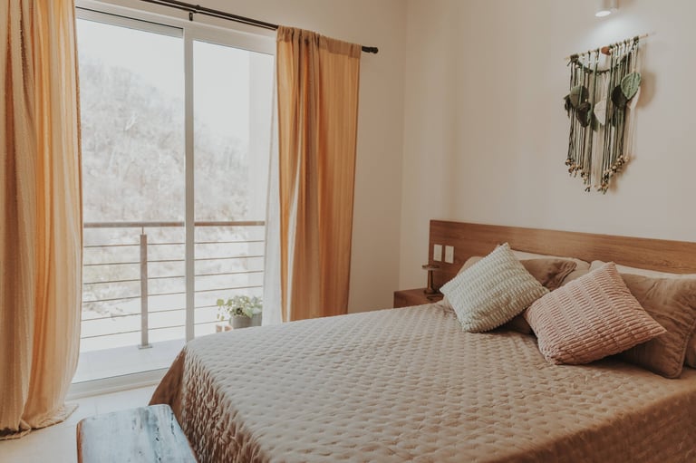 Modern bedroom with queen bed, decorative pillows, large window with orange curtains, and macrame wall art