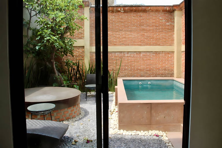 View through glass doors of two outdoor courtyards with a round wooden hot tub on left and a rectangular blue swimming pool on right, surrounded by brick walls and gravel